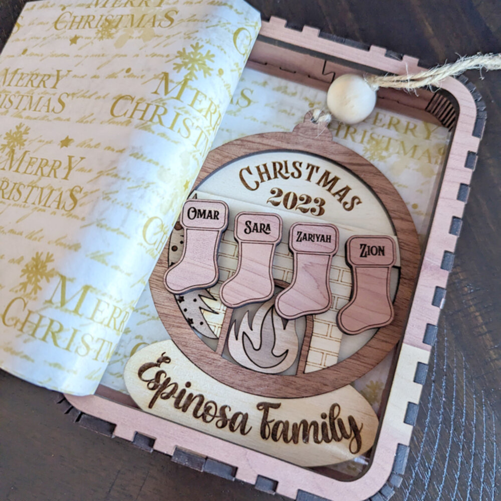 A layered wooden ornament made from a variety of woods sites in a cedar wooden box.  Cedar stockings hang over a maple and walnut fireplace, all framed in a wooden snow globe with the family name underneath.