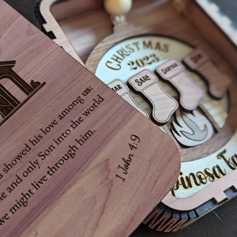 A layered wooden ornament made from a variety of woods sites in a cedar wooden box.  Cedar stockings hang over a maple and walnut fireplace, all framed in a wooden snow globe with the family name underneath.