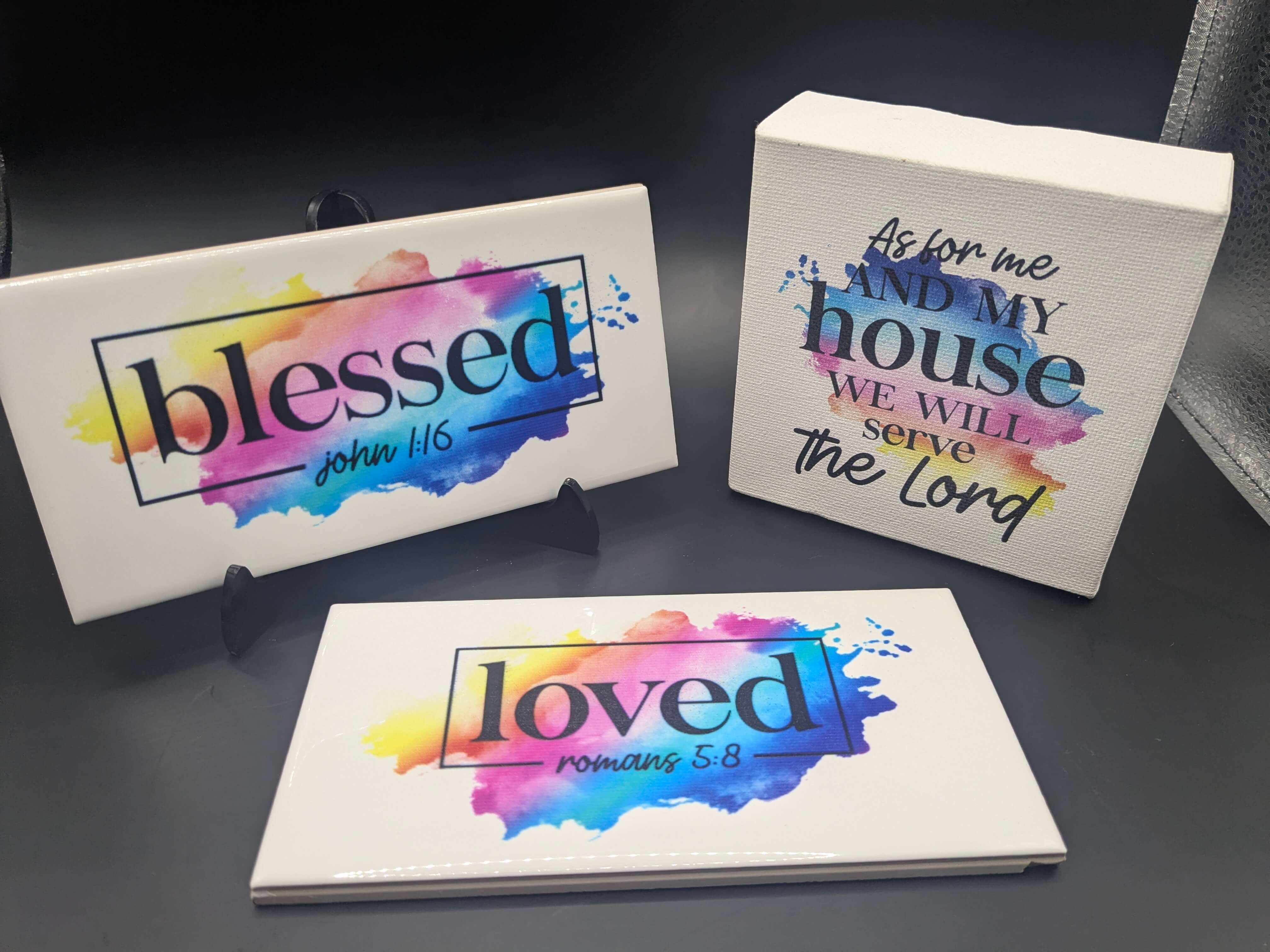 Colorful UV-printed Christian home décor signs featuring Bible verses: ‘Blessed – John 1:16,’ ‘Loved – Romans 5:8,’ and ‘As for me and my house we will serve the Lord’ on ceramic tiles and a canvas block with vibrant watercolor backgrounds.