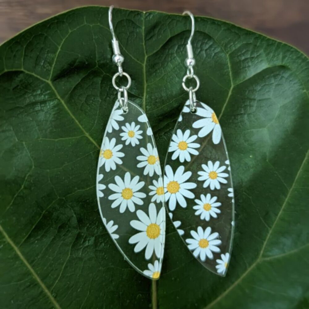 clear earrings with printed daisies hang in front of a green leaf