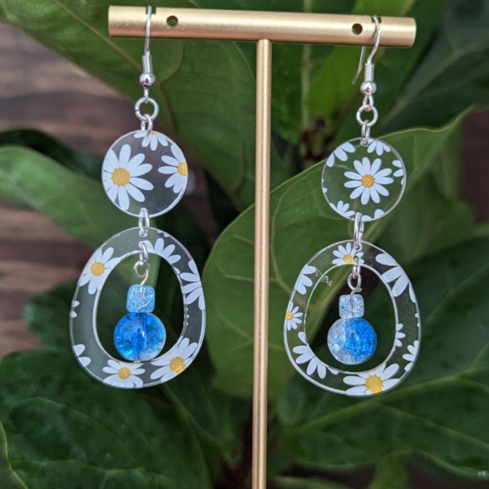 Circular daisies with blue bead earrings hang from an earring stand in front of a green leaf