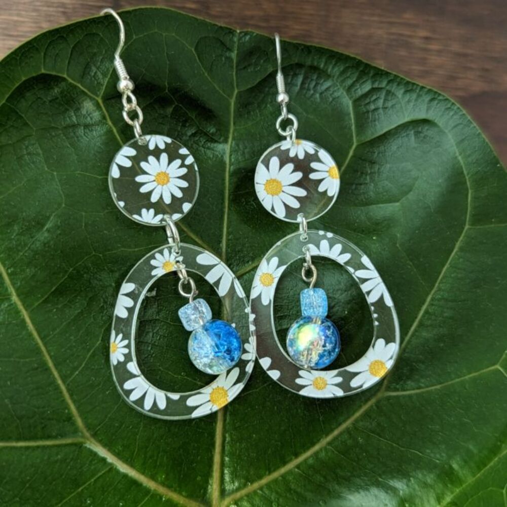 Circular daisies with blue bead earrings sit on a green leaf
