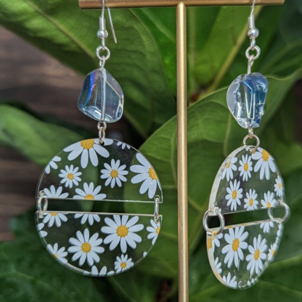 Daisy half circles earrings with bead hanging in front of green foliage