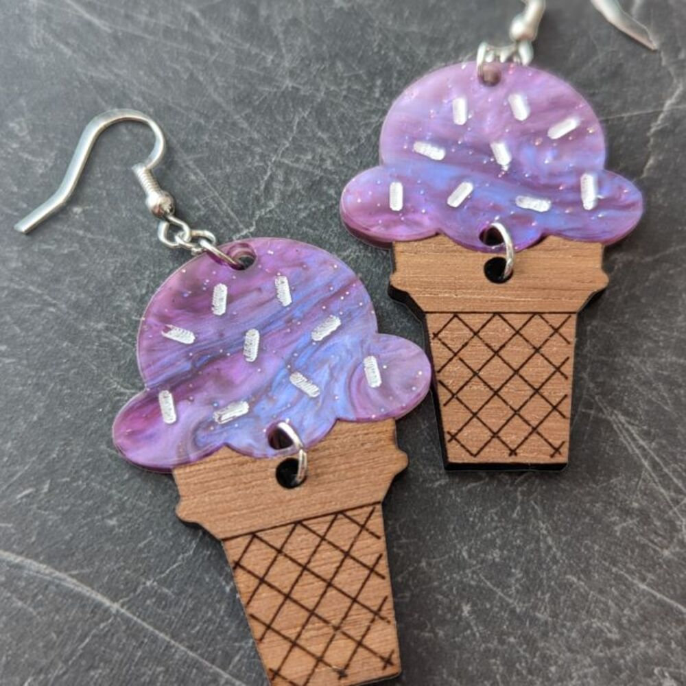 Dangle earrings featuring purple acrylic "ice cream with sprinkles" attached to an engraved walnut "cone" against a dark granite