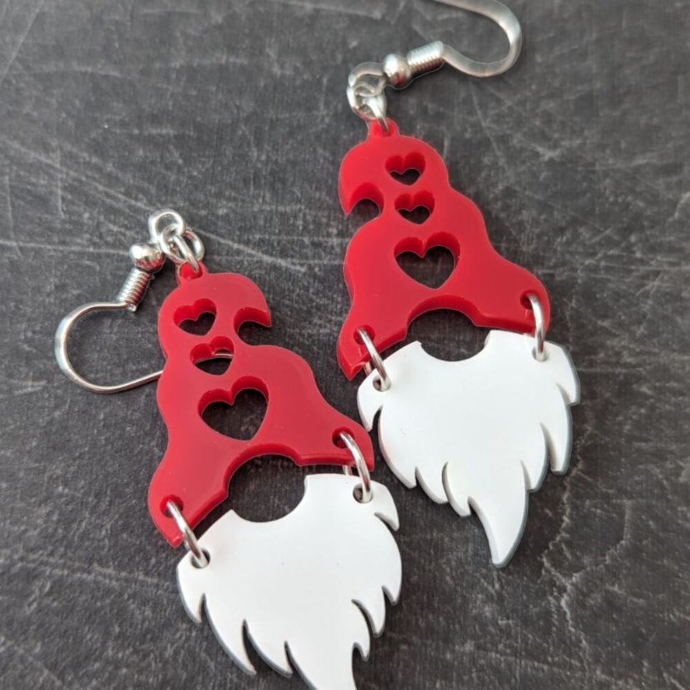 A pair of earrings with a white beard and red hat, with hearts cut out of the hat; against a dark gray background