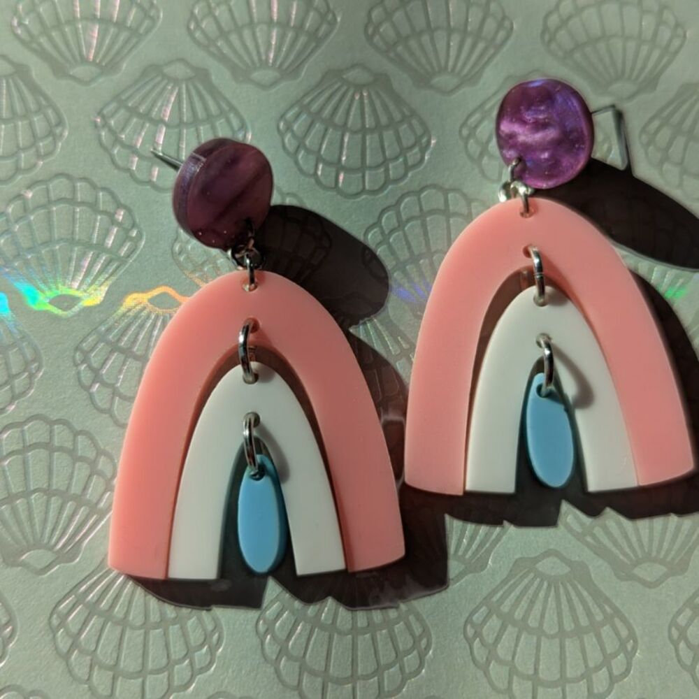 A pair of purple stud earrings dangling a pink rainbow, dangling a white rainbow, dangling a blue rainbow; against a shell background with light refracting across it