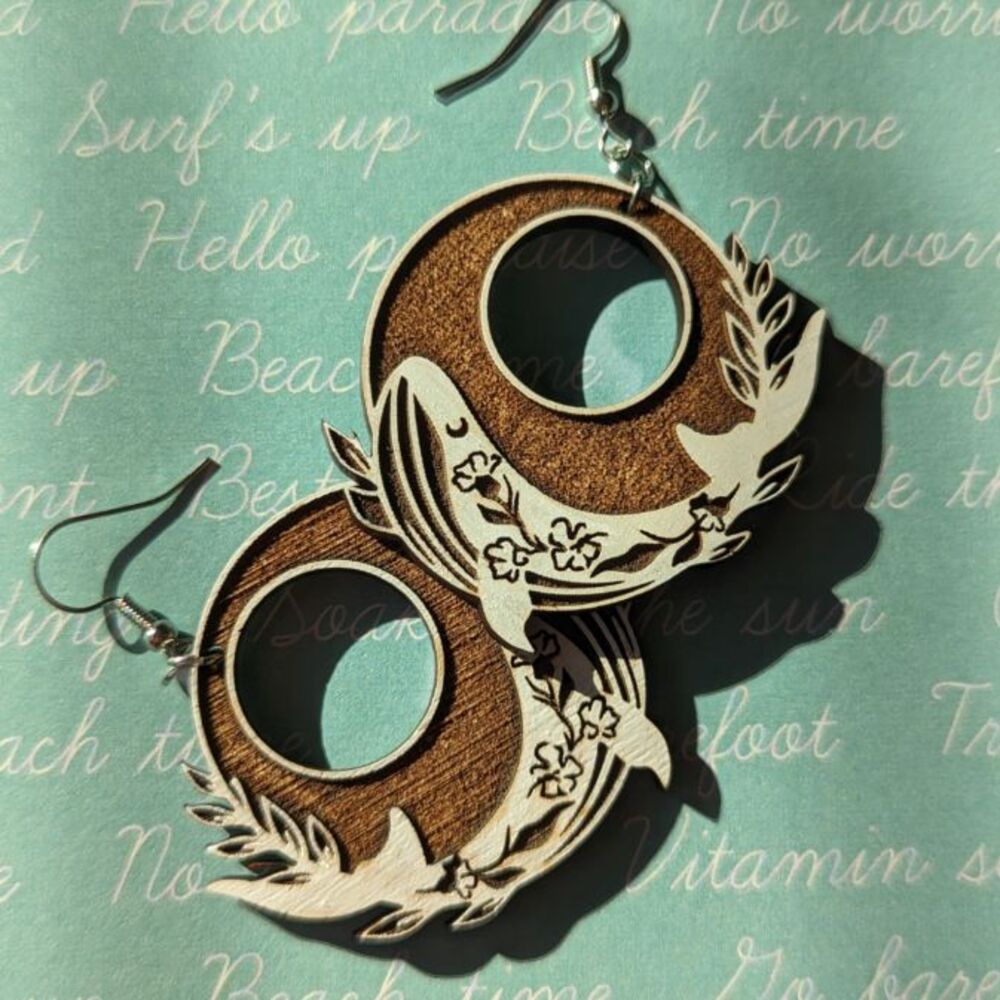 A pair of circular dangle earrings with painted white whales, against a surf's up background