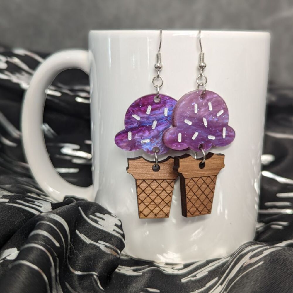 Dangle earrings featuring purple acrylic "ice cream with sprinkles" attached to an engraved walnut "cone" hanging from a white porcelain mug