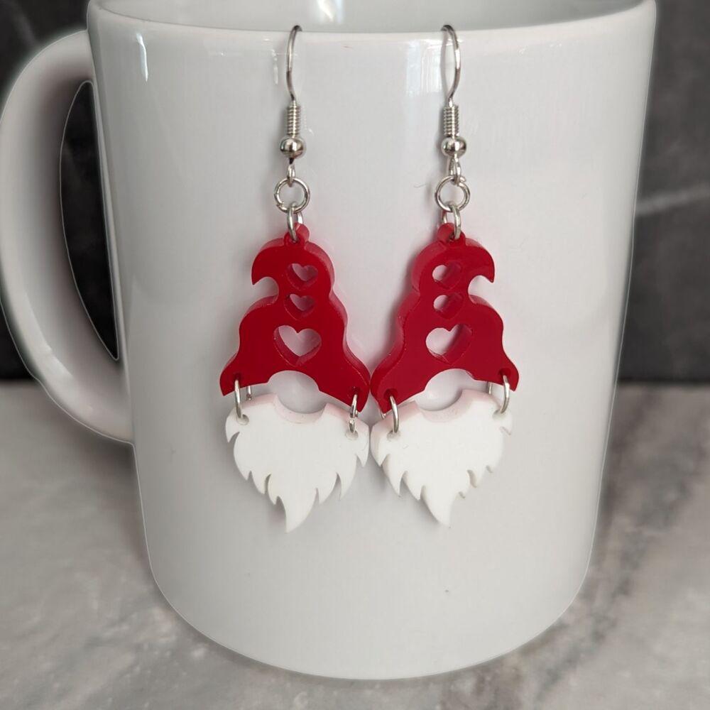 A pair of earrings with a white beard and red hat, with hearts cut out of the hat; hanging from a white porcelain mug
