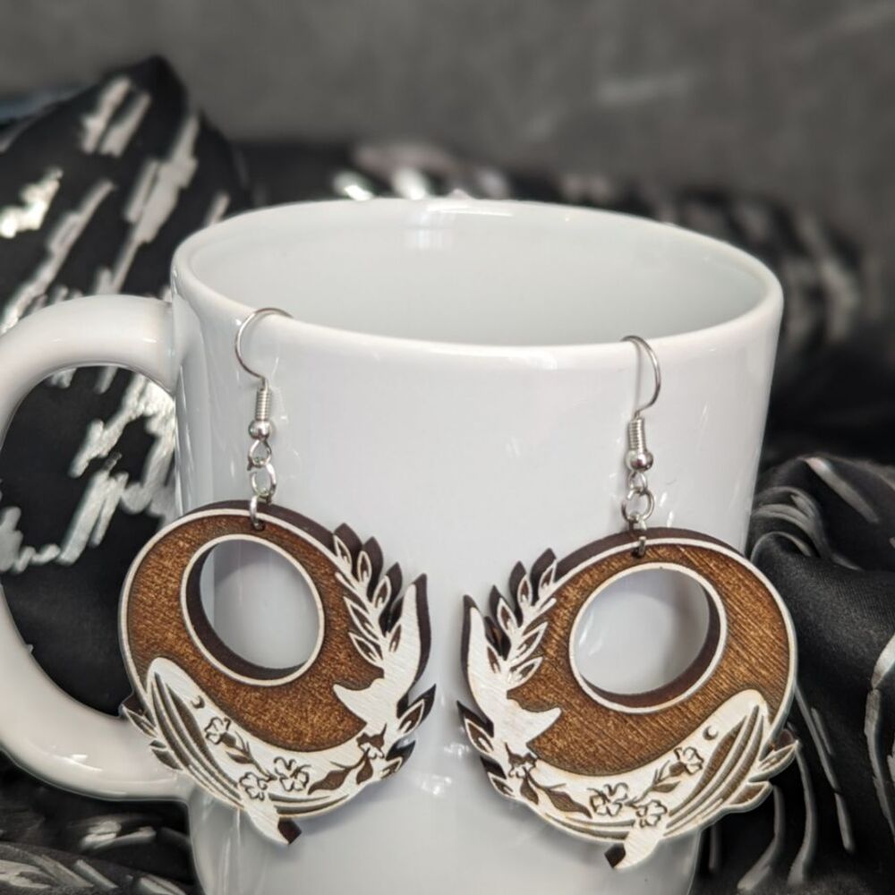 A pair of circular dangle earrings with painted white whales, against a white porcelain mug