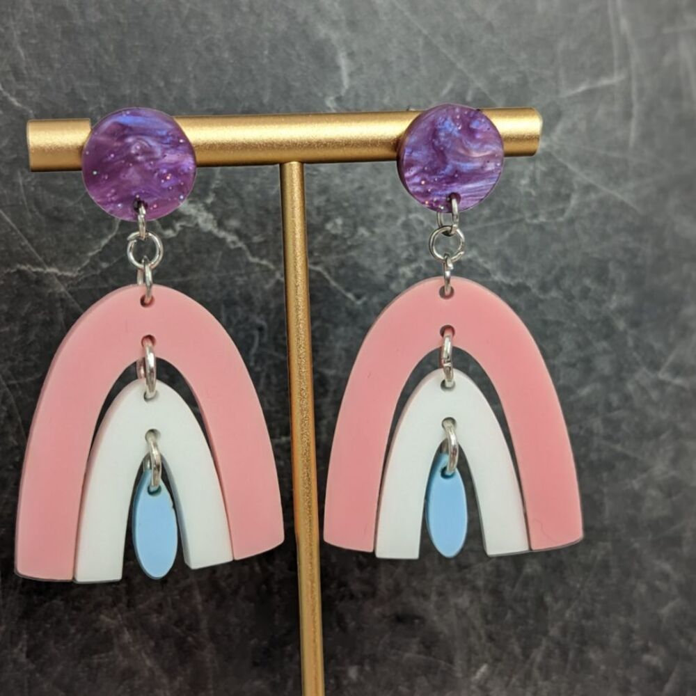 A pair of purple stud earrings dangling a pink rainbow, dangling a white rainbow, dangling a blue rainbow