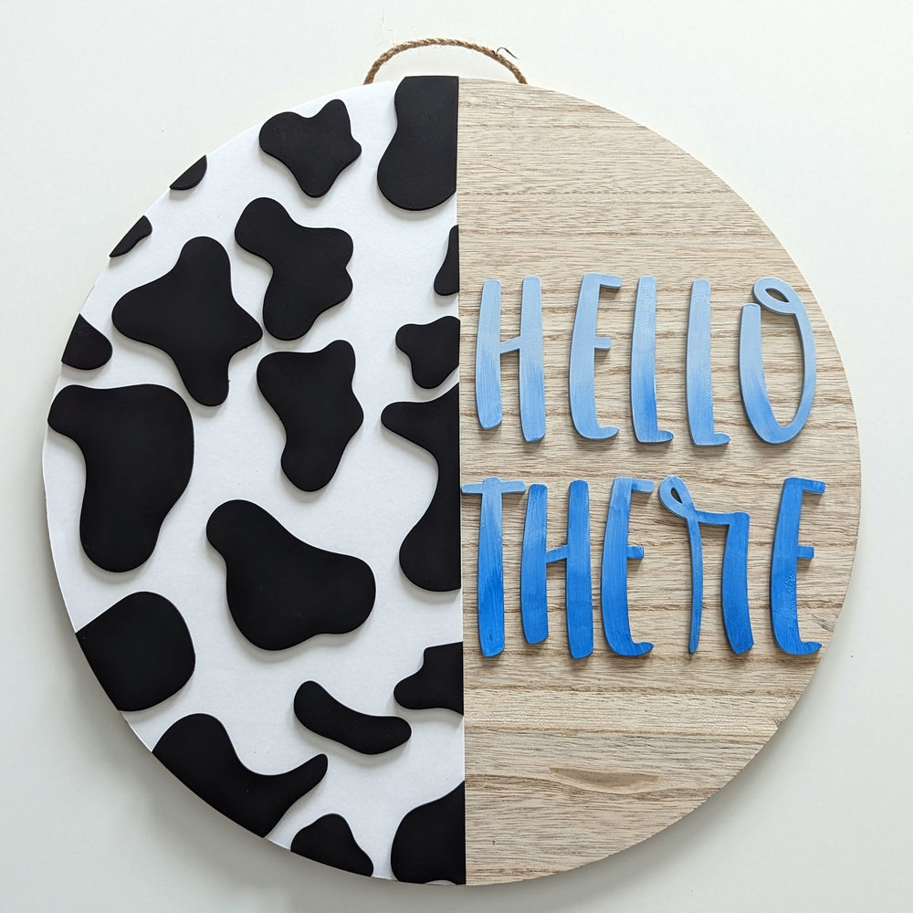 A round sign with jute hanger. The left hand side has cow print, the right says "Hello There"
