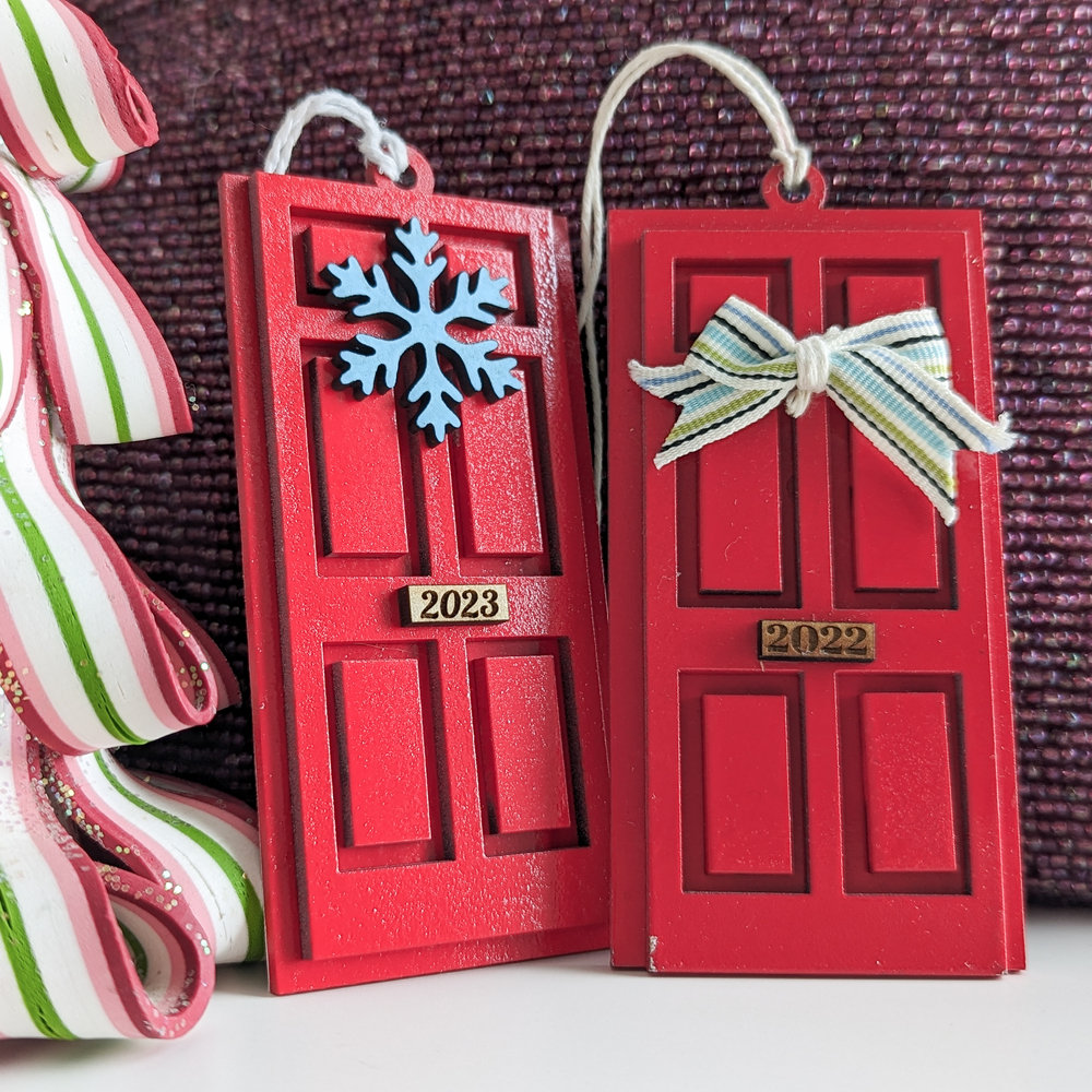 Two ornaments in the shape of a door, painted red, one with a snowflake wreath. the other with a bow.