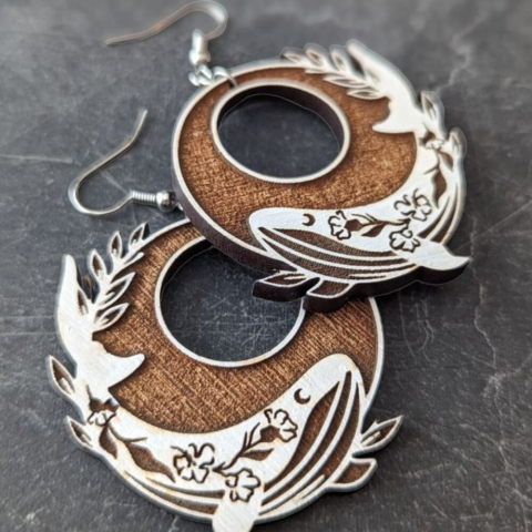 A pair of circular dangle earrings with painted white whales, against a dark gray background