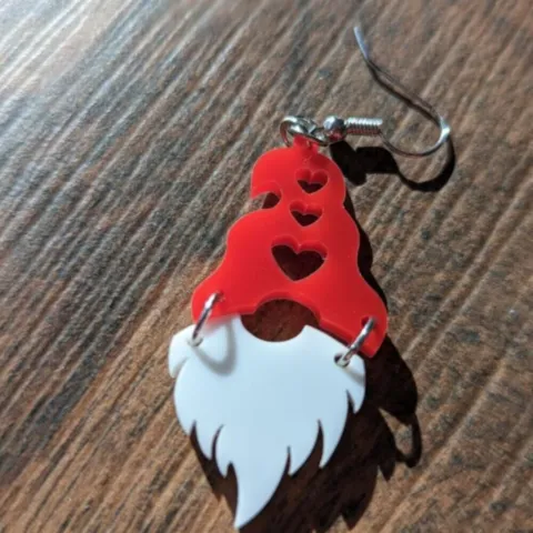 A single earrings with a white beard and red hat, with hearts cut out of the hat; against a wood background