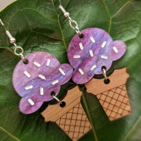 Dangle earrings featuring purple acrylic "ice cream with sprinkles" attached to an engraved walnut "cone" against a green leaf