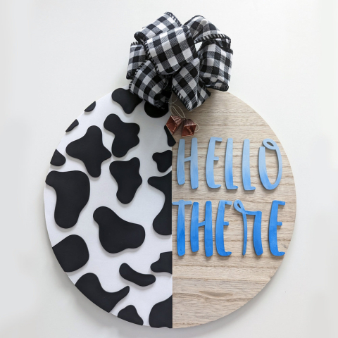A round sign with jute hanger. The left hand side has cow print, the right says "Hello There"