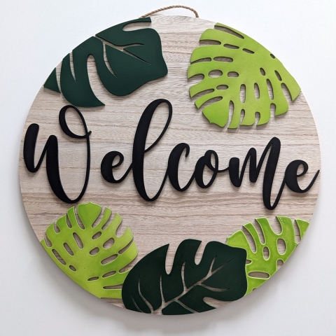 A round sign with the word Welcome across it. The sign also has a number of large tropical looking green leaves and a jute hanger.