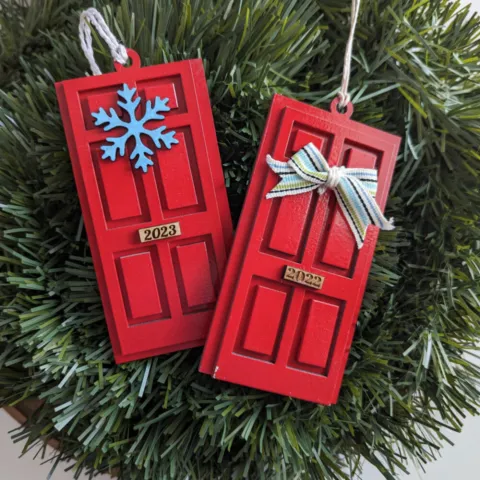 Two ornaments in the shape of a door, painted red, one with a snowflake wreath. the other with a bow.