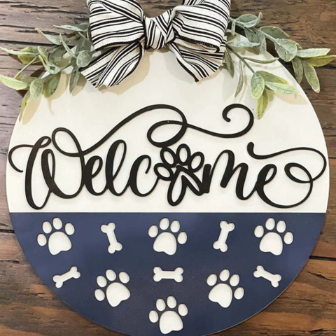 A round sign with a cursive "Welcome" across the middle. The "o" of the welcome is a paw shape. The bottom 1/3 of the sign is a contrasting color and has paw print and dog bone shapes cut out of it. The sign is topped with a beautiful bow.
