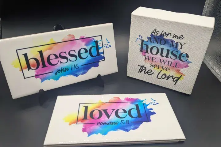 Colorful UV-printed Christian home décor signs featuring Bible verses: ‘Blessed – John 1:16,’ ‘Loved – Romans 5:8,’ and ‘As for me and my house we will serve the Lord’ on ceramic tiles and a canvas block with vibrant watercolor backgrounds.